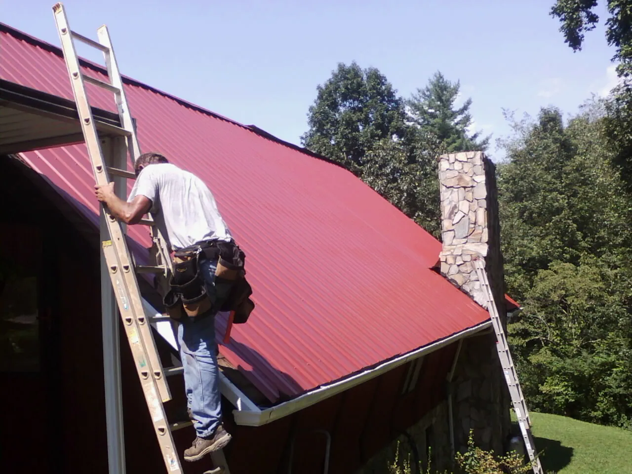 Roofer on a ladder beside a red metal roof and stone chimney — Soffit & Fascia Repair by Roofing Brothers in Leland